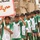 A group of young boys wearing matching green and white soccer uniforms with Iraqi flag patches are lined up, likely for a sports event or parade. One boy holds a sign with Arabic text reading 'حي المعامل' (Al-Maamel neighborhood). A military armored vehicle is visible in the background, suggesting this event takes place in a military-adjacent or secured area.