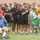 A group of male football/soccer players of various ages are lined up on a grass field, likely during a pre-match ceremony such as a national anthem. Players are wearing multiple team kits including black, red, green, and yellow uniforms. A man in a wheelchair wearing a blue sweatshirt is positioned at the front-left, and a formally dressed adult male stands at the far right.