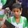 A young boy wearing a white and green soccer jersey with the number 11 and what appears to be an Iraqi flag patch is holding a dark folder/certificate and a plastic bag with items including goalkeeper gloves. He appears to be at a youth soccer event or award ceremony. His expression is serious or contemplative.
