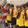 A U.S. Army soldier wearing an 82nd Airborne Division patch is interacting with a group of young boys dressed in soccer uniforms, appearing to sign autographs or distribute items. The children are lined up in yellow and black soccer jerseys with visible numbers, suggesting a youth soccer event or community outreach activity. A second soldier is visible in the background near a chain-link fence.