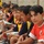 A row of young boys in sports uniforms are seated along a chain-link fence, likely on the sideline of a soccer field. The boys appear to be players waiting their turn, wearing various team jerseys in red, black, yellow, and navy colors. The foreground boy in a red jersey looks directly at the camera while others look forward.