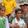 A U.S. Army soldier wearing ACU (Army Combat Uniform) poses with two young boys at what appears to be a soccer stadium. The soldier has his arms around both children, one of whom is holding a soccer ball. The boys are wearing green and white soccer jerseys with 'PEAK' branding and Iraqi flag patches.
