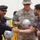 A U.S. Army soldier identified as McGuire participates in what appears to be a ceremonial exchange of soccer balls with a local official wearing a dark uniform and beret. A civilian holding a microphone shakes hands with the uniformed local official while the soldier holds a soccer ball in the background. The setting suggests a community engagement or goodwill event.