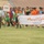 A youth football (soccer) tournament opening ceremony or parade is taking place on a grass field inside a stadium. Participants carrying Arabic-language banners and signs are marching across the pitch, representing different teams or districts. The crowd includes children and adults wearing various colored sports kits.