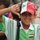 A young boy poses energetically for the camera, making a finger-frame gesture with both hands and smiling broadly. He is wearing a green and white striped sports jersey with an Iraqi flag patch on the chest, along with a multicolored cap featuring the Iraqi flag colors (red, white, black, and green). Several other children are visible in the background.