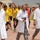 Three individuals wearing white medical coats over military camouflage uniforms walk across a stadium track, with the center figure visibly wearing a stethoscope. Behind them, a large group of athletes in yellow sports jerseys with Arabic text celebrate or march together. The setting appears to be a sports event or ceremony held at an outdoor stadium.