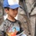 A young boy wearing a blue and white baseball cap with Arabic script and a graphic t-shirt featuring a soccer player with the number 8 is holding a package of what appears to be sports clothing or a soccer kit. In the background, a person in military camouflage uniform with a weapon slung over their shoulder is partially visible. The setting suggests a humanitarian or community outreach interaction between military personnel and a local child.