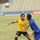 A football (soccer) match is taking place in a stadium with largely empty stands. Two players are competing for the ball — one wearing a yellow Adidas kit with Arabic text, and another in a blue kit. The ball is airborne above them as they jostle for position.