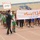 A youth sports parade or opening ceremony is taking place at a stadium, with groups of young athletes in various football/soccer uniforms marching on a running track. Participants are carrying Arabic-language banners and signs identifying their teams or districts. The large central banner reads 'قاطع (9) نيسان - اللجنة الرياضية' (District 9 Nissan - Sports Committee), suggesting a local or regional youth sports tournament in Iraq.