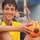 A young man wearing a yellow and blue Adidas soccer jersey is posing with an orange soccer ball branded 'Laser Galit'. He is smiling directly at the camera with wet hair, suggesting recent physical activity. In the background, individuals in military camouflage uniforms are partially visible.