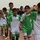 A group of young boys wearing green and white PEAK-branded soccer uniforms with Iraqi flag patches are walking together on a running track, smiling and waving at the camera. The jerseys display numbers including 5 and 11. The setting appears to be a sports stadium or athletic facility.