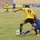 A soccer match is being played in a stadium with sparse seating. The focal point is a physical challenge between a player in yellow (number 19) and a player in blue near the ball. Additional players and what appears to be a referee are visible in the background.