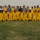 A large group photo of a soccer team wearing yellow and black uniforms posing on a grass field inside a stadium. Several individuals in military camouflage uniforms and caps are integrated into the back row of the group. Two young boys in civilian clothes are kneeling at the front left of the photo.
