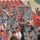 A crowd of people in a stadium with red seating are celebrating enthusiastically. Many individuals are wearing blue-grey camouflage military uniforms, mixed with civilians in casual clothing. The group appears to be celebrating a sporting event victory, spraying water bottles and waving yellow flags.