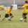 A soccer match is being played on a grass field inside a stadium with blue seating visible in the background. Three players are actively engaged near the ball: one in a blue uniform and two in yellow and black uniforms, one of whom wears number 17. Arabic-language advertising banners line the perimeter boards, suggesting the match is taking place in an Arabic-speaking country.