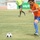 A soccer match is in progress on a grass field, with a player in an orange jersey and blue shorts dribbling toward a black-and-white soccer ball. A referee in a yellow-green jersey is visible in the background, along with at least one other player. Advertising banners with Arabic text line the background perimeter.