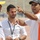 Three men are engaged in a conversation at what appears to be a sports stadium or athletic track. The man on the right, wearing a white and orange shirt and a gray Adidas cap, is pointing and appears to be giving directions or instructions. The setting suggests a sporting event or training session.