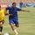 A soccer match is taking place in a stadium with advertising boards visible in the background. Two players are prominently featured in the foreground, one in a blue kit and one in a yellow kit with Arabic script, competing for the ball. Additional players and what appears to be a referee or coach in yellow are visible in the background.