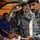 The image shows a senior military or police officer in blue digital camouflage uniform speaking to individuals seated on a sports bench under a dugout canopy. Several uniformed personnel are present, including one photographing the scene with a DSLR camera. The setting appears to be a sports facility, likely a football/soccer stadium.