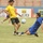 A soccer match is in progress on a grass field, with a player in a blue uniform executing a sliding tackle against a player in a yellow jersey who is controlling the ball. Advertising banners in Arabic and English are visible along the sideline in the background. A third player in blue is partially visible in the background.