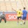 A soccer match is being played in a stadium with empty stands. Three players are visible near the goal: a goalkeeper wearing a gray 'ARMY' shirt and gloves, a player in yellow jersey number 9, and a player in a blue uniform. The scene appears to be a casual or training match based on the informal goalkeeper attire.