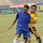 Two male soccer players are competing for possession of the ball during a match played on a grass field. One player wears a blue Puma kit with white socks, while the other wears a yellow and black kit. Advertising boards including 'Zain' and 'Showai' are visible in the background along stadium seating.