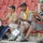 Three men are seated in the stands of a stadium with red seats, playing musical instruments as fans or supporters. The central figure plays a brass trumpet while wearing a yellow bandana around his neck, and nearby are two drums including a blue bass drum labeled 'AL-SADAKA'. The scene appears to be a sporting event where these musicians are providing live percussion and brass accompaniment to support their team.