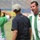 Three men are gathered on a football pitch, likely during a training session or pre-match activity. The man on the right is wearing a Real Betis green-and-white striped jersey and appears to be speaking or gesturing. The man on the left, wearing a number 12 PEAK-branded green jersey, is holding a small camera or device.