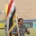 A uniformed military or police officer carries the Iraqi national flag along a stadium running track. He is accompanied by at least one other similarly uniformed individual behind him. The setting appears to be a formal ceremony or parade held inside a sports stadium.