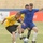 A soccer match is being played on a grass field inside a stadium with empty yellow seats in the background. Two primary players are competing for the ball — one in a yellow jersey with black shorts and another in a full blue kit. Advertising banners with Arabic text are visible along the sideline.