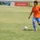 A male soccer player wearing an orange Valencia CF jersey with number 7 and blue shorts is dribbling a soccer ball on a grass field. The player appears to be in mid-stride, approaching the ball during what looks like a match or training session. Advertising banners, including one for 'Zain', are visible along the stadium perimeter in the background.
