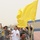 A group of uniformed personnel, wearing blue-grey digital camouflage uniforms with visible shoulder patches, are celebrating on what appears to be an athletic track at a stadium. A man in the foreground is kneeling and waving a large bright yellow flag while smiling broadly. The scene suggests a post-competition victory celebration.