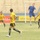 A soccer match is being played on a grass field in a stadium. One player in a blue jersey is airborne, appearing to head the ball, while several opposing players in yellow and black uniforms surround him. The stadium has empty tan/beige concrete bleachers in the background with Arabic-language advertising banners along the sideline.