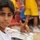 A young boy wearing a white and orange sports jersey poses for the camera while holding a signed soccer ball and a water bottle. He is making a peace/victory sign with his fingers and appears to be at a sporting event or stadium. The background shows red stadium seats and other individuals wearing yellow uniforms.
