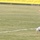 A young athlete wearing a white uniform is performing a handstand or cartwheel on a grass football/soccer field. The uniform appears to have 'IRAQ' written on the shorts, suggesting this may be a national team player celebrating or warming up. Arabic-language advertising banners and a 'Zain' telecom logo are visible in the background.