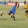 A soccer match is in progress on a grass field, with one player in a blue and yellow uniform competing against two players wearing yellow and black uniforms. The central player appears to be attempting to control or pass the ball mid-action. Advertising banners including 'SHOWC' and 'LCD' are visible along the sideline in the background.