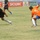 A soccer match is being played on a grass field with players from two opposing teams — one in black uniforms and one in orange and blue uniforms. The central action shows a player in black dribbling the ball while a player wearing number 5 in orange attempts to challenge or intercept. Advertising banners for 'Shownic', 'LCD', and 'Zain' are visible along the sideline in the background.