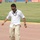 A man dressed in casual attire — white button-up shirt, khaki pants, sandals, and sunglasses — is kicking a soccer ball on a running track at a stadium. In the background, players in orange and blue uniforms are visible on the grass field. Sponsor banners including 'ZAIN' and 'SHOWNIC' are visible along the perimeter.