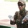 A person in military/tactical gear is holding two plastic water bottles, one in each hand. They are wearing a desert camouflage cap, dark sunglasses, a green balaclava/neck gaiter, and a camouflage uniform with a unit patch on the right sleeve. The individual appears to be smiling and standing near a green container in an outdoor setting.