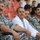 A group of men in blue digital camouflage uniforms with Iraqi flag patches are seated in what appears to be a stadium with red seats. Among them sits one man wearing a white lab coat or medical coat, suggesting he may be a medical officer or civilian. The group appears to be observing an event taking place off-camera.