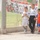 Three young boys are walking together near a chain-link fence at what appears to be a football/soccer facility. Two of the boys are wearing white jerseys with 'IRAQ' printed on them and Iraqi flag patches, while the third wears a red and blue FC Barcelona-style jersey. A large banner featuring soccer players and Arabic text is displayed on the fence behind them.