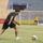 A soccer match or training session is taking place in a stadium with largely empty blue seats. A player in a black uniform is the focal point, leaning forward and approaching a soccer ball on a grass field. Additional players and what appears to be a referee or official in yellow are visible in the background.