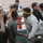 A crowd of young males gathered in what appears to be a stadium or sports facility. A man wearing a yellow headband and white shirt is leaning toward a younger boy in a green shirt, appearing to interact closely with him. Several onlookers surround them, with one person capturing the moment on a mobile phone.