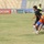 A soccer match is being played in a stadium with sparse spectator seating. Two players are engaged in a physical challenge near the center of the pitch, with one player in an orange and blue kit appearing to fall while contesting the ball with a player in a black kit. A referee in a yellow-green jersey and another player in black are visible in the background.