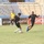 A football (soccer) match is being played near a goal on a grass pitch. Two outfield players in black uniforms are competing for the ball close to the penalty area, while a goalkeeper in a yellow jersey stands in position behind them. The stadium appears largely empty, with blue and beige seating visible in the background.