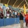 A crowded sports stadium scene showing a large group of spectators packed into the stands behind a blue barrier wall. Several individuals in military camouflage uniforms and one person in a white medical coat are positioned at the field level, appearing to manage or monitor the crowd. Yellow and orange flags are being waved by fans in an energetic atmosphere.