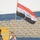 A uniformed military or police officer carries the Iraqi flag on a wooden pole while walking on a red athletic track inside a stadium. The flag displays the distinctive red, white, and black horizontal stripes with green Arabic script in the center. The stadium bleachers are visible in the background, appearing largely empty.