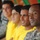 A group of individuals are seated together, appearing to watch an event. The foreground features a U.S. Army soldier in ACU (Army Combat Uniform) camouflage, seated alongside several civilians wearing yellow shirts. The image has a shallow depth of field, keeping the soldier in sharp focus while the background figures are progressively blurred.