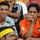 A group of young male football fans are seated in stadium stands, watching a match. Two prominent figures in the foreground wear football jerseys — one in a white and green kit and another in an orange Valencia CF jersey with the LFP (La Liga) patch. The fans display mixed emotions, with one smiling and another appearing tense or anxious.