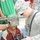 A U.S. Army soldier in ACU (Army Combat Uniform) is showing a professional video camera to a smiling young child wearing a camouflage cap and a maroon shirt with the number 8. The interaction appears friendly and positive, suggesting a community engagement or humanitarian outreach activity. The setting appears to be an outdoor public area with other people visible in the background.